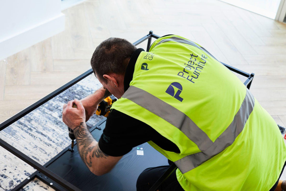 Worker in a high visibility vest kneels indoors while assembling a black metal frame over a dark panel, with tools and a patterned rug nearby.