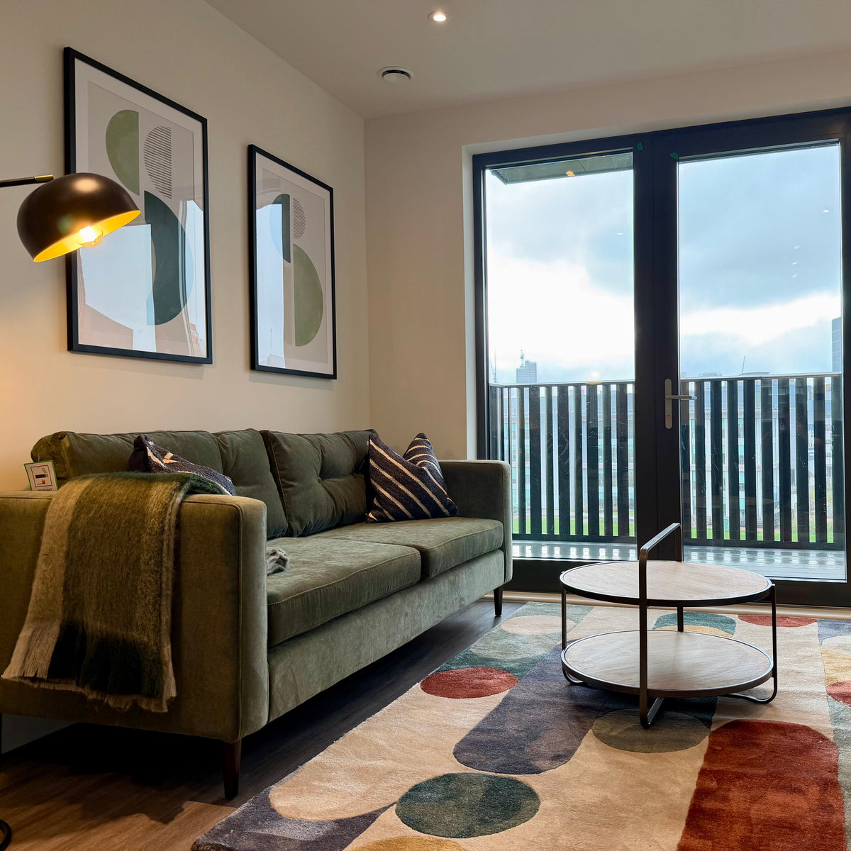 Modern living room with a green sofa and blanket beside a floor lamp and framed art overlooking a balcony through glass doors with a patterned rug and coffee table