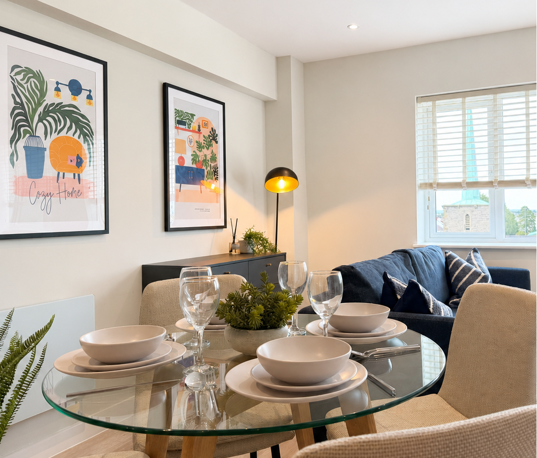 Bright dining room with a round glass table set with bowls and wine glasses surrounded by beige chairs beside a navy sofa framed botanical artwork and a window
