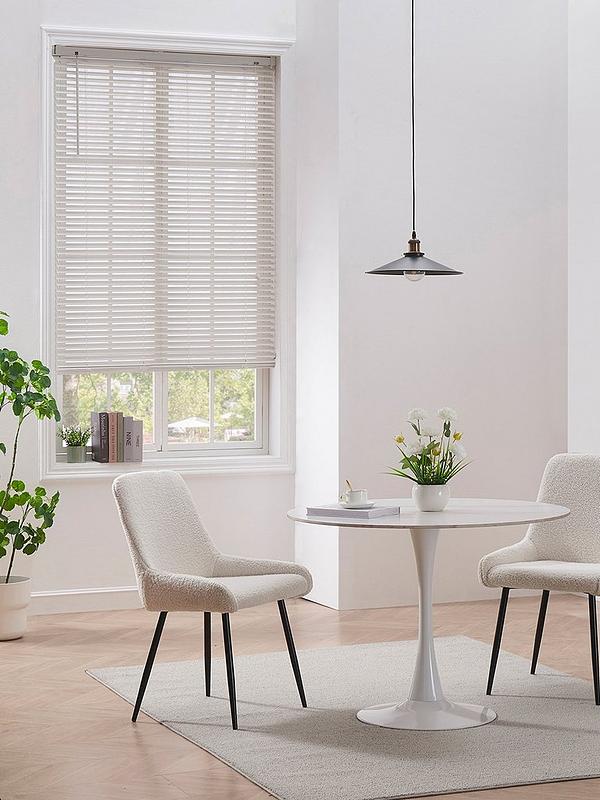 Bright minimalist dining area with a round white table, upholstered chairs, pendant light, window blinds, potted plants, books, and a neutral rug over wood flooring