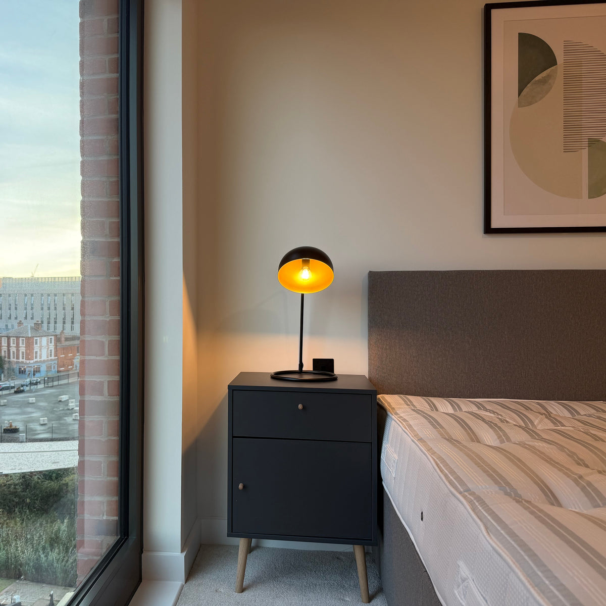 Modern bedroom with a dark bedside table, glowing round lamp, upholstered headboard, striped bedding, framed wall art, and a city view through the window