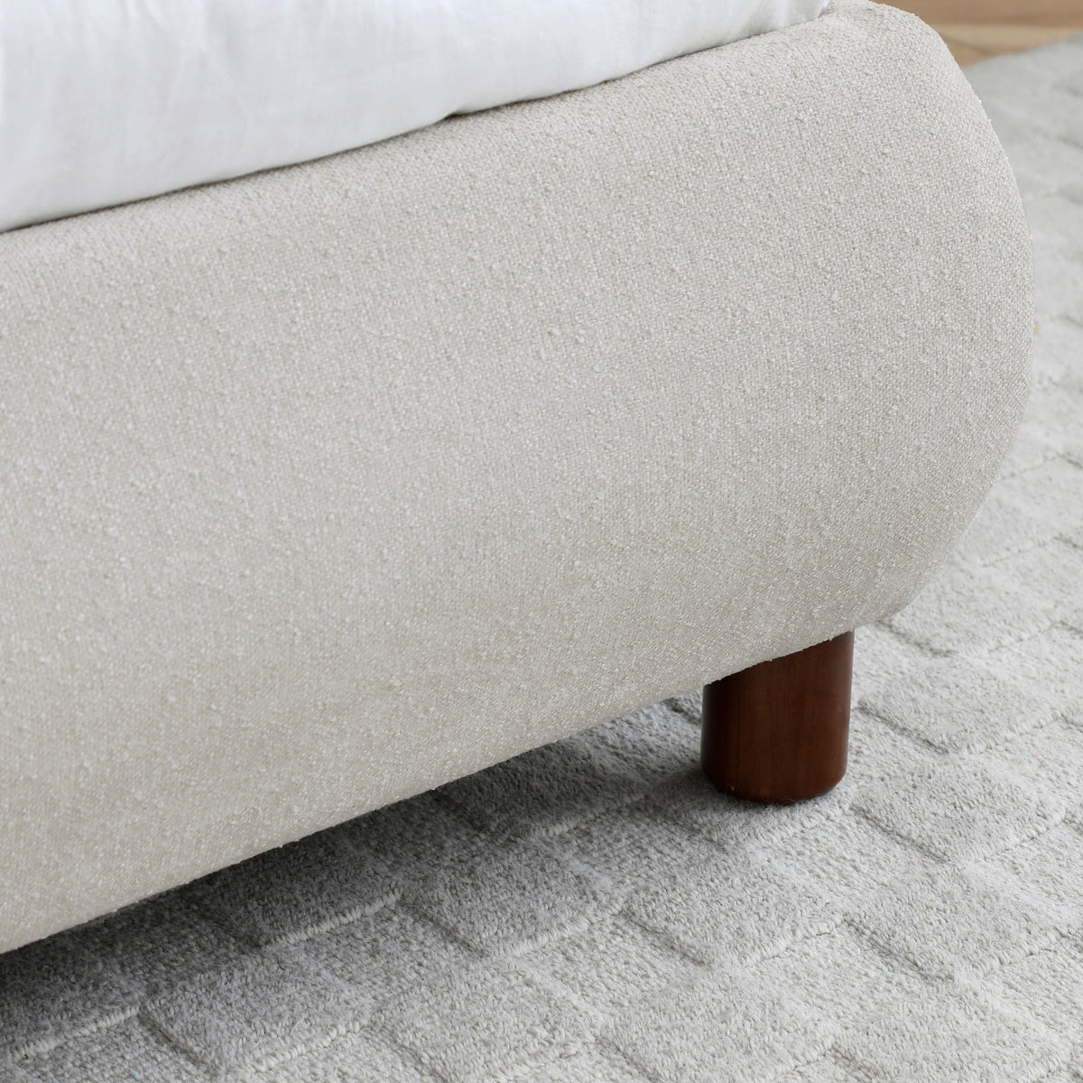 Close up of a cream upholstered bed frame with a rounded edge and cylindrical wooden leg resting on a light textured rug beside white bedding